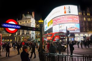 Vista de plaza londinense por la noche con pantallas grandes, la imagen del metro y mucha gente